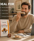 A man in a beige sweater sits at a desk drinking from a glass cup of coffee while a bag of 10Café Carri Delicio Ginseng coffee pods and a single pod are displayed in the foreground. The image includes marketing text on the left side.