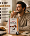 A man in a beige sweater sits at a wooden table holding a cup of coffee, with a box of 101CAFFE Betagi Cioccolata chocolate coffee capsules placed in front of him. The scene includes promotional text on the left highlighting rich chocolate indulgence, instant energy, protein and cocoa goodness, and creamy smooth taste.